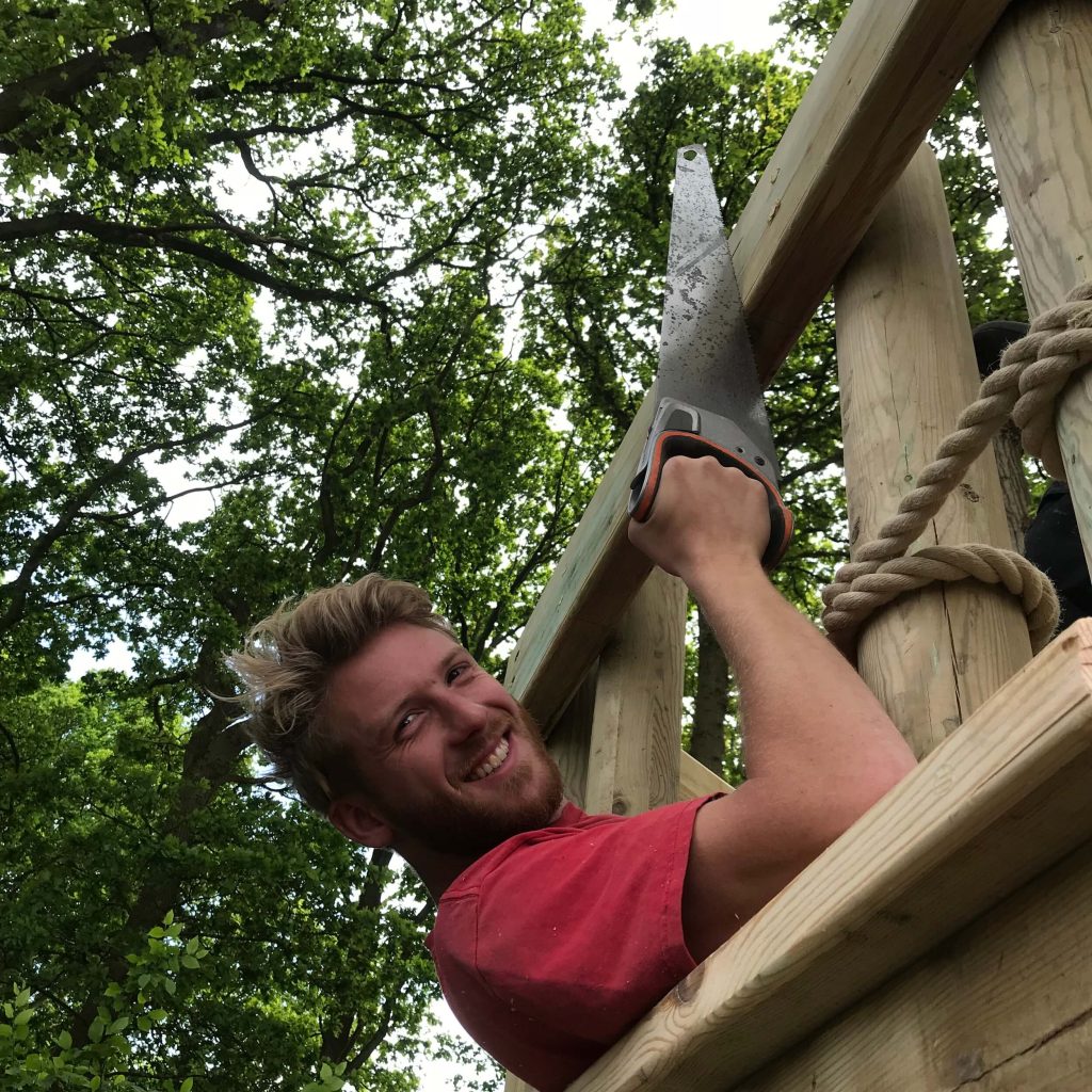 Man in red T-shirt and holding a hand saw laying on a Treehouse Deck Platform, smiling and about to cut a balustrade timber.