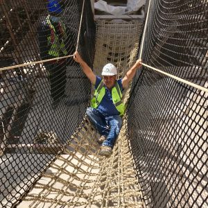 Rope Bridge builder person sat within a Net Rope Bridge with natural colour Scramble Cargo Net floor and black safety netting sides, holding on to structural super rope.
