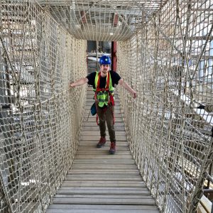 Rope Bridge builder person standing in centre of Rope Bridge wearing harness and safety equipment, surrounded by safety netting to sides and above head, standing on timber Rope Bridge slats.