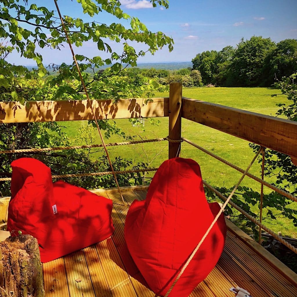 A suspended tree platform bathed in sunshine with a view overlooking green fields in Effingham, Surrey. The deck has two large red comfy bean bags.