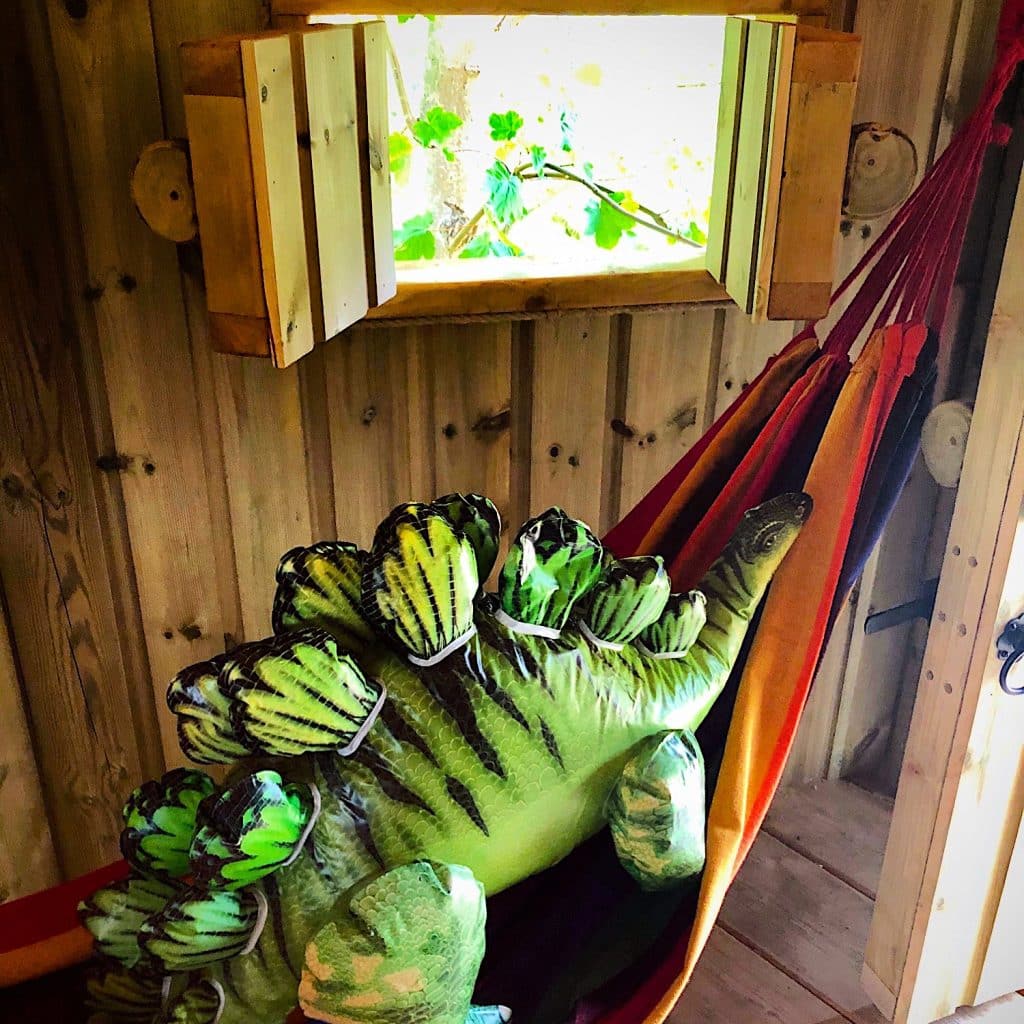 A bright rainbow hammock hanging inside a treehouse under an open treehouse window. The hammock has an inflatable green dinosoar in it.