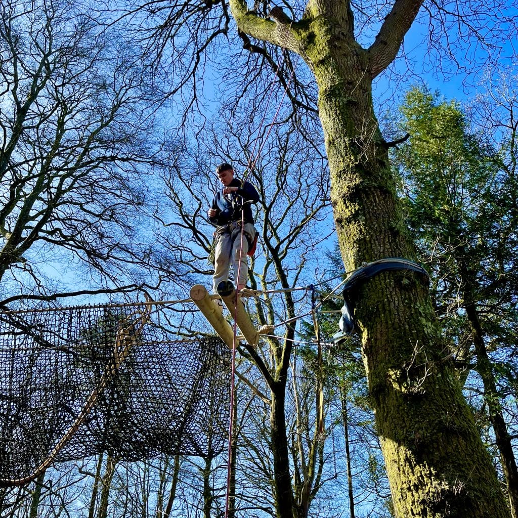 A man stood at the end of a partially constructed rope bridge installation in the Lake District. he balances on two beams of wood while being secured to a tree trunk by a rope harness.