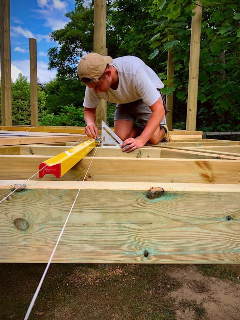 A man constructing the base level of a garden treehouse. He is using a spirit level and string to measure and space the timber beams.