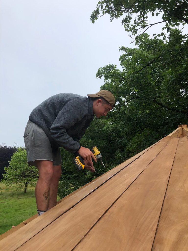 A man drilling a timber beam into the roof of a treehouse.