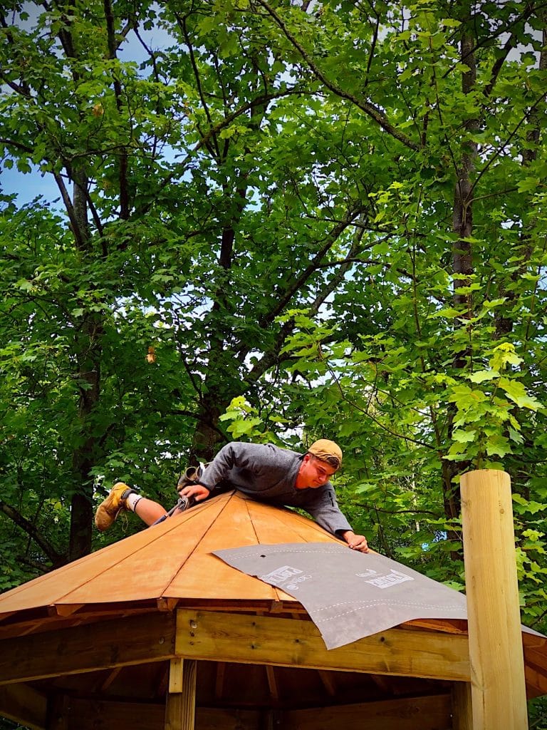 A man balancing on top of a treehouse roof in Surrey, UK, holding a sheet of felt to be secured onto the roof to waterproof it.