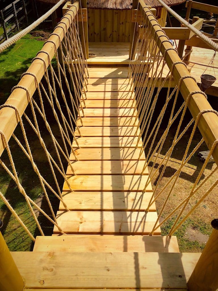 A rope bridge between a large family treehouse and long garden slide.
