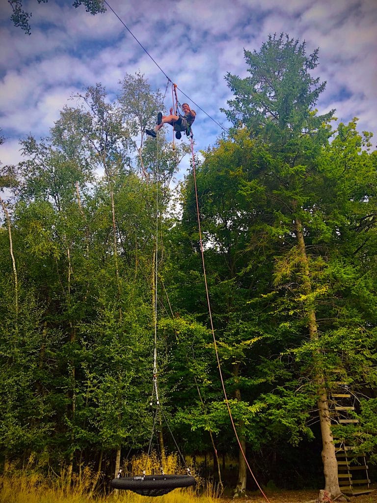 A man suspended high in the air between two giant fir trees. The man is hoisting up a nest swing on a harness and rope.