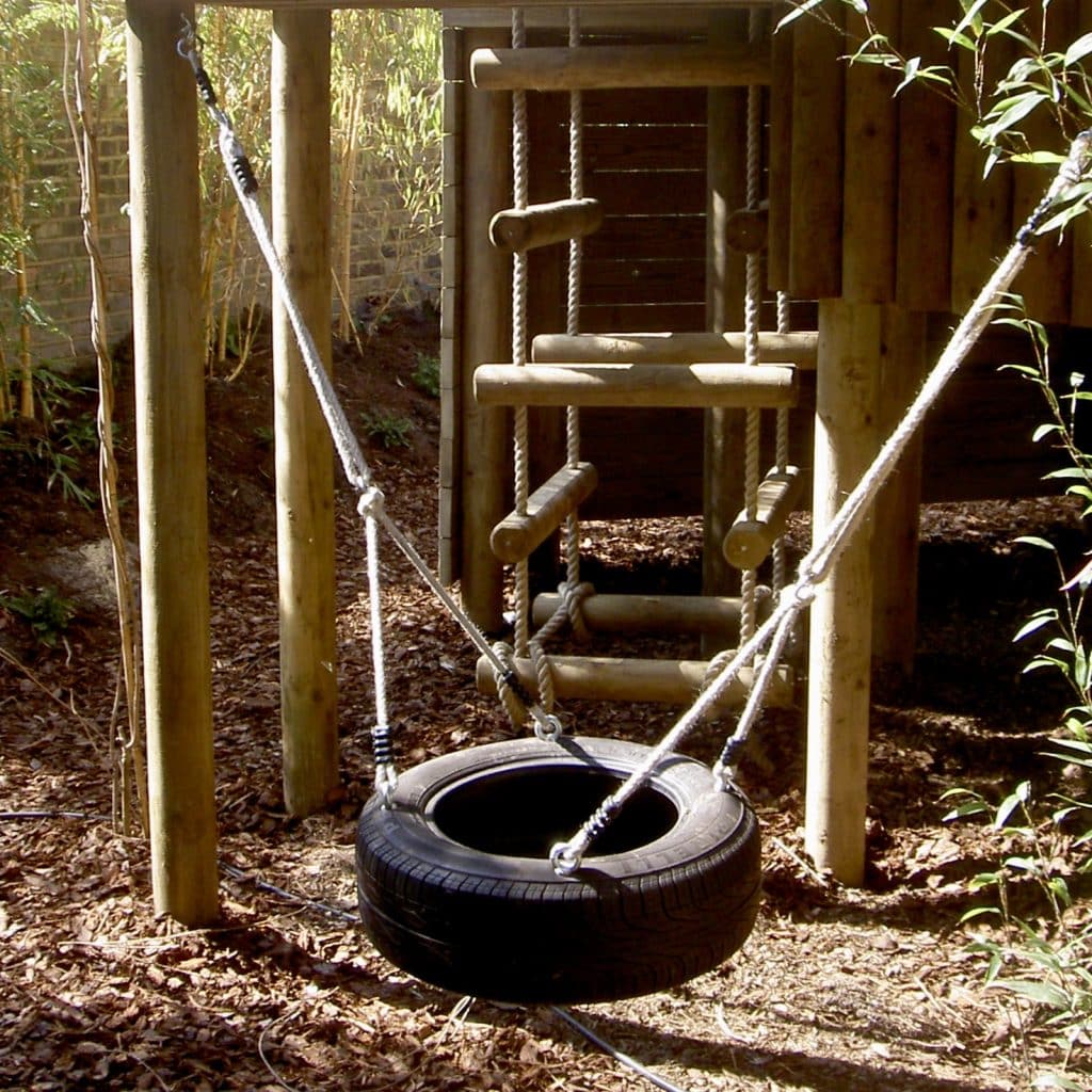 A large black tyre swing suspended under a deck of a family garden treehouse. There are four wooden pillars and a chunky four sided rope ladder behind the swing. The floor is barked.