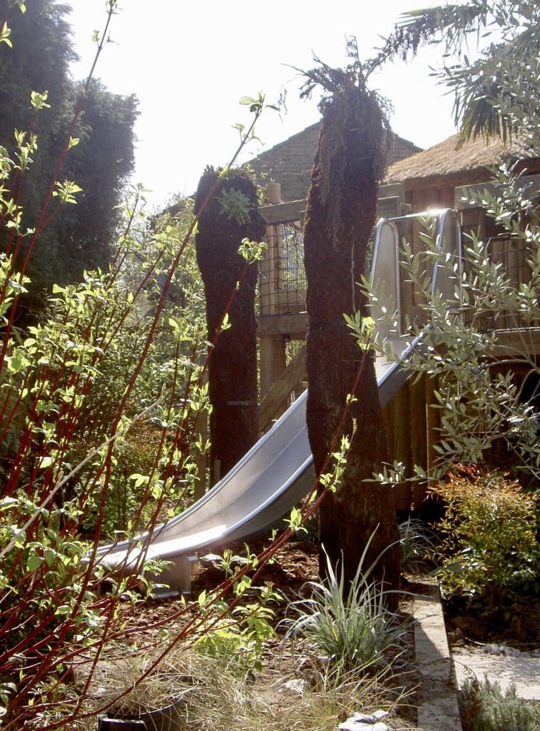 A shiny stainless steel 2 metre slide leading down from a landscaped garden treehouse in Windsor, UK.
