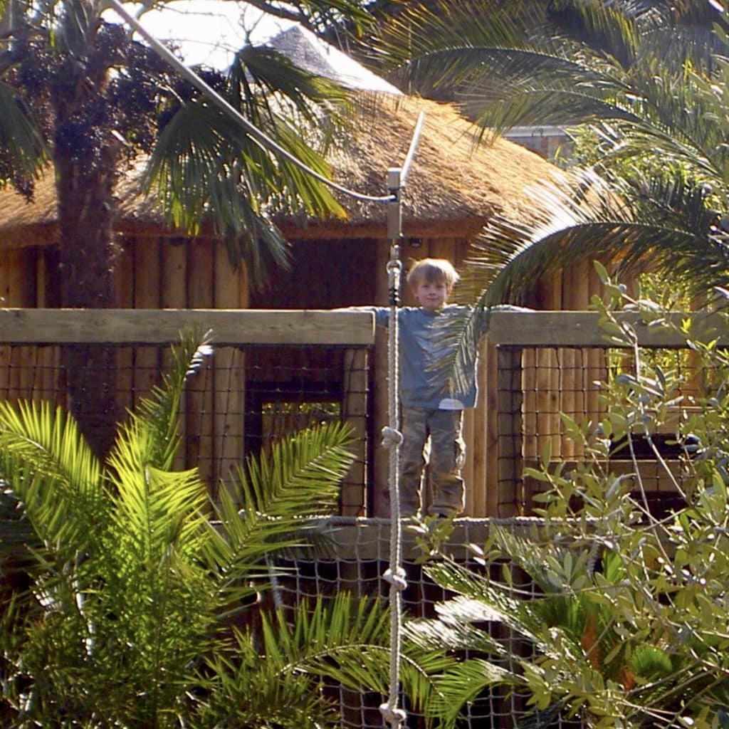 A small boy standing at the deck of a garden zip wire, in front of a large family treehouse.