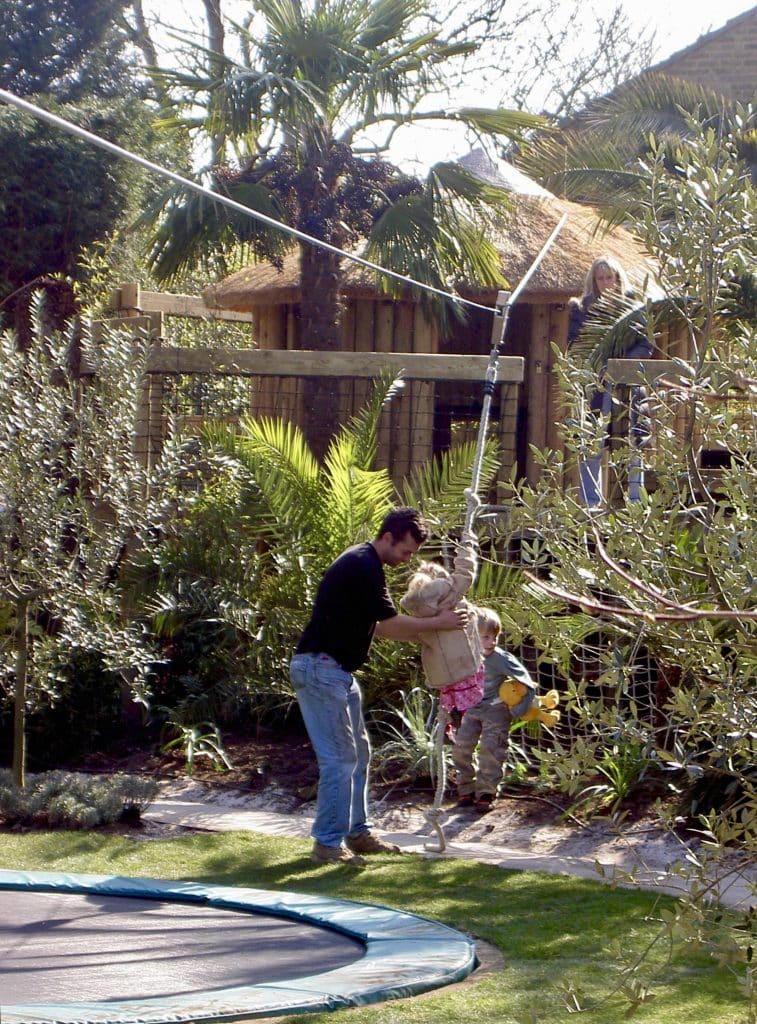 A father helps his small daughter to sit on their garden zip slide as the young brother and his mum watch on. The mum stands on the tree deck of their luxury garden treehouse. zip slide
