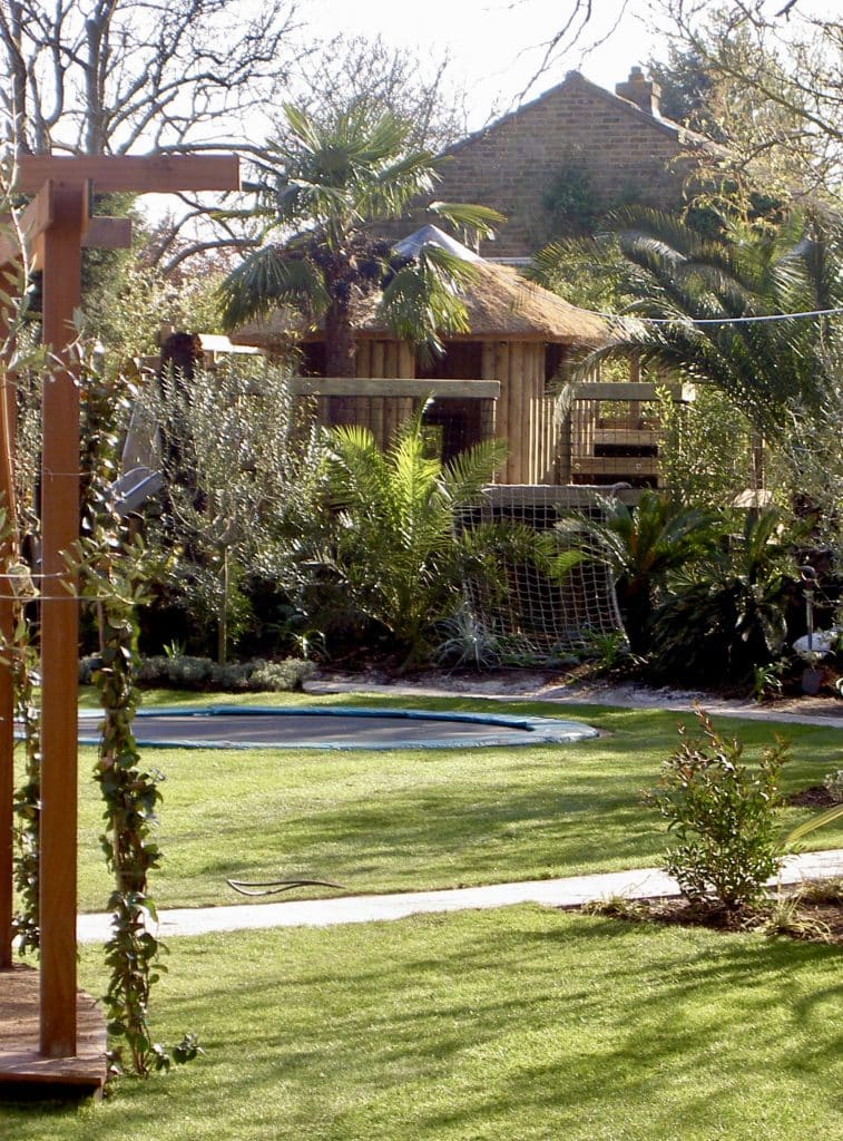 A lawned garden with a sunken trampoline leading to a landscaped garden treehouse surrounded by palm trees and tropical shrubbery.