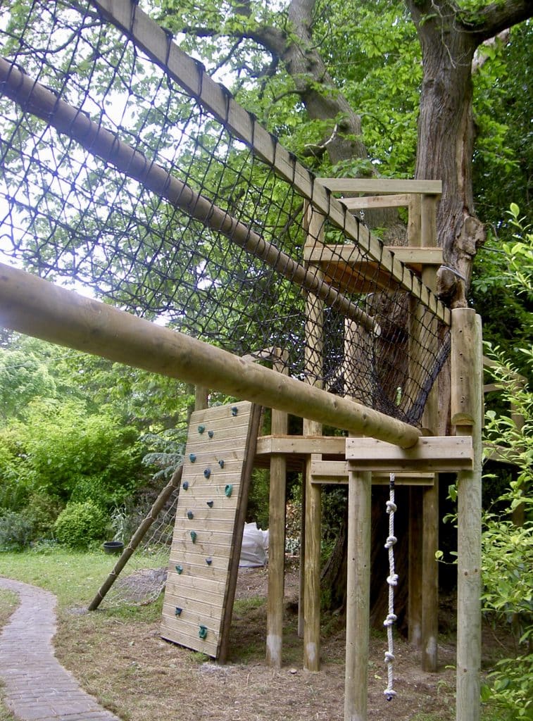 A sturdy rope bridge across a cobbled footpath leading to a further adventure play area built around a large oak tree in a private garden. The play area features a timber climbing wall, a climbing rope, a double height tree deck with three separate platforms and a large cargo scramble net. The adventure play area is surrounded by large green and lush trees.