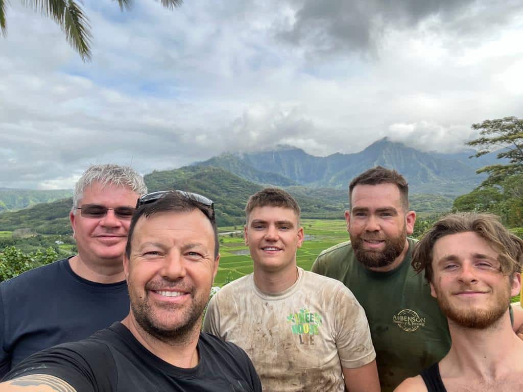 Paul, Charlie and the Treehouse Life team of master carpenters arrive on Kauai Island to begin their commercial project to build 4 rope bridges at a luxury private resort. The team are smiling, with a mountain in the background, and palm trees to the side of them.